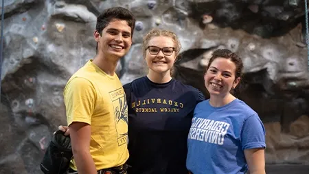 Three students smiling in front of climbing wall