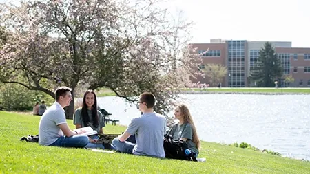 Four students reading while sitting on grass by lake