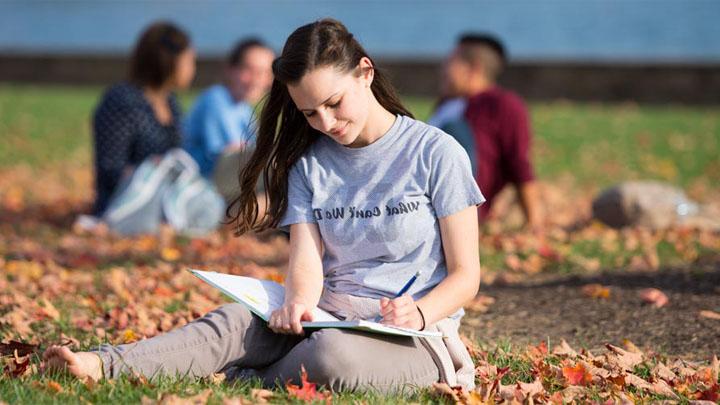female student sitting in grass in the fall writing in a notebook